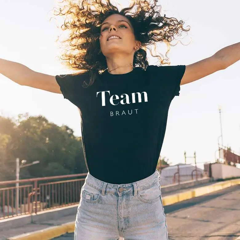 Frau in einem schwarzen "Team Braut"-T-Shirt, strahlend mit offenen Armen unter freiem Himmel. Perfekt für Junggesellinnenabschiede.